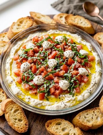 A glass dish of layered bruschetta dip with fresh tomatoes basil crumbled cheese and a drizzle of olive oil surrounded by toasted baguette slices