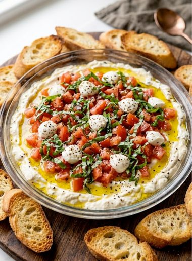A glass dish of layered bruschetta dip with fresh tomatoes basil crumbled cheese and a drizzle of olive oil surrounded by toasted baguette slices