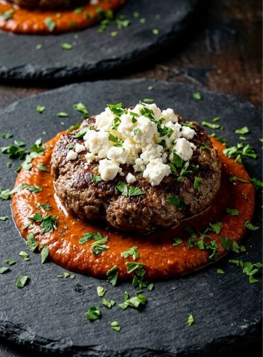 A juicy keto burger patty served over creamy tomato sauce and topped with crumbled feta cheese and fresh parsley on a dark plate