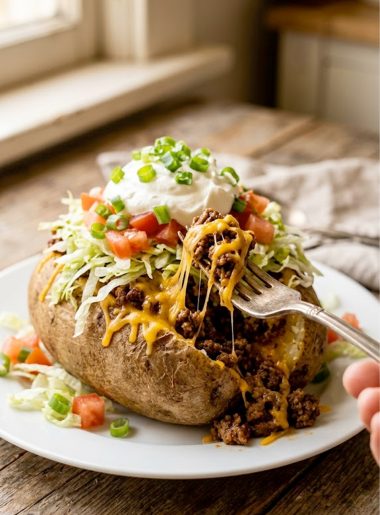 A large russet baked potato split open and loaded with seasoned taco meat, melted cheddar cheese, shredded lettuce, diced tomatoes, sour cream and sliced green onions on a white plate