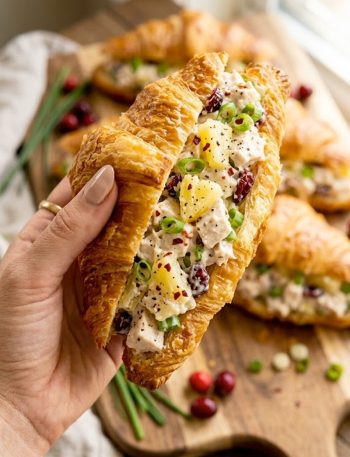 A hand holding a buttery flaky croissant sandwich generously filled with creamy Maui Mama chicken salad with visible pineapple pieces, dried cranberries, and red pepper flakes