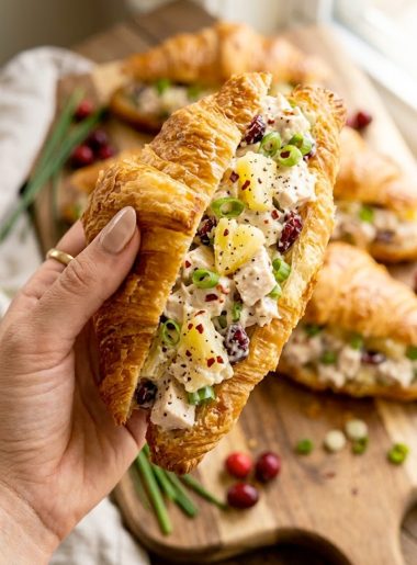 A hand holding a buttery flaky croissant sandwich generously filled with creamy Maui Mama chicken salad with visible pineapple pieces, dried cranberries, and red pepper flakes