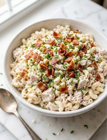 A large white bowl piled high with creamy chicken pasta salad topped with crispy bacon crumbles and fresh chopped chives on a marble surface