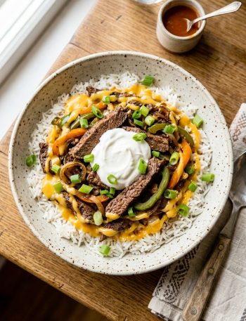 High protein Philly cheesesteak bowl with tender seasoned beef strips sauteed peppers and onions over fluffy rice drizzled with glossy melted cheddar cheese sauce and topped with sour cream and spring onions in a speckled ceramic bowl