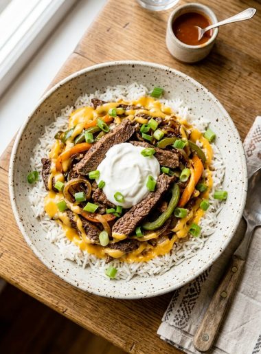 High protein Philly cheesesteak bowl with tender seasoned beef strips sauteed peppers and onions over fluffy rice drizzled with glossy melted cheddar cheese sauce and topped with sour cream and spring onions in a speckled ceramic bowl