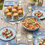 A spread of picnic-style cold meals on a wooden outdoor table including mini sliders, pasta salad, and caprese skewers