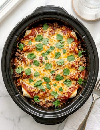 A slow cooker beef enchilada casserole with layers of seasoned ground beef, flour tortillas, melted cheddar cheese, and rich red enchilada sauce served in a black slow cooker