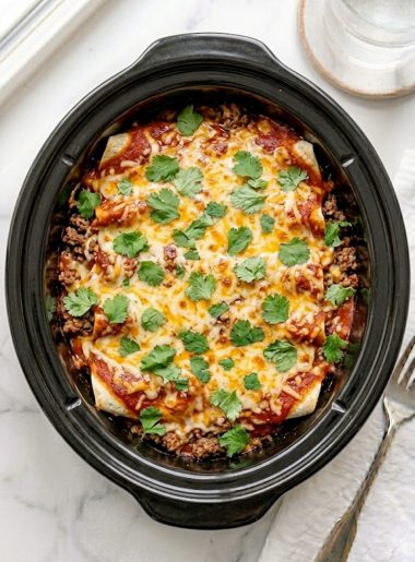 A slow cooker beef enchilada casserole with layers of seasoned ground beef, flour tortillas, melted cheddar cheese, and rich red enchilada sauce served in a black slow cooker