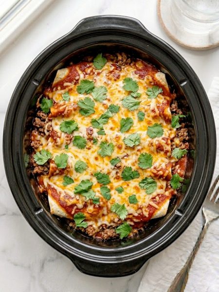 A slow cooker beef enchilada casserole with layers of seasoned ground beef, flour tortillas, melted cheddar cheese, and rich red enchilada sauce served in a black slow cooker