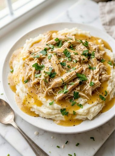 A white plate piled with tender shredded chicken and rich golden gravy served over creamy mashed potatoes garnished with fresh parsley