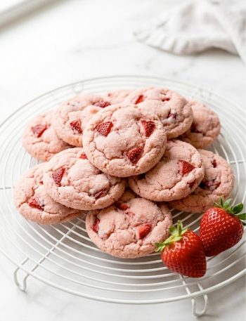 A pile of soft and chewy strawberry cookies with a pink sugary exterior on a white cooling rack with fresh strawberries beside them