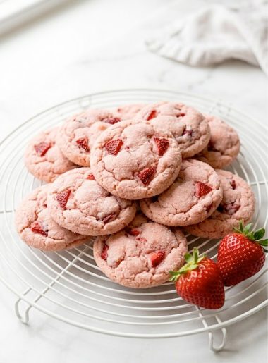 A pile of soft and chewy strawberry cookies with a pink sugary exterior on a white cooling rack with fresh strawberries beside them