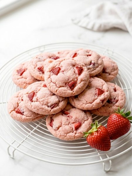 A pile of soft and chewy strawberry cookies with a pink sugary exterior on a white cooling rack with fresh strawberries beside them