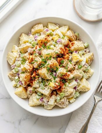 A large white ceramic bowl of sweet Amish potato salad made with tender Yukon Gold potato chunks, diced red onion, green onion, celery, and hard-boiled eggs coated in a rich creamy sweet dressing topped with smoked paprika and fresh chives on a white marble surface