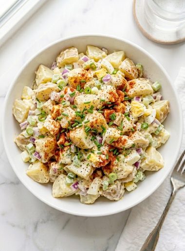 A large white ceramic bowl of sweet Amish potato salad made with tender Yukon Gold potato chunks, diced red onion, green onion, celery, and hard-boiled eggs coated in a rich creamy sweet dressing topped with smoked paprika and fresh chives on a white marble surface