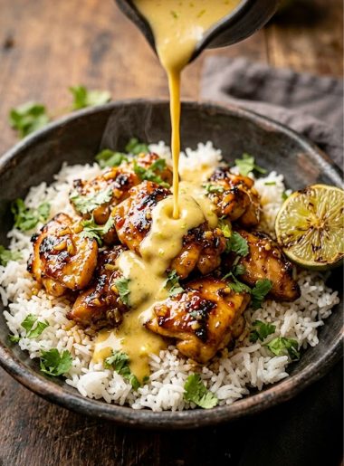 A bowl of sweet chili chicken over jasmine rice with creamy coconut lime sauce being drizzled on top and fresh cilantro garnish