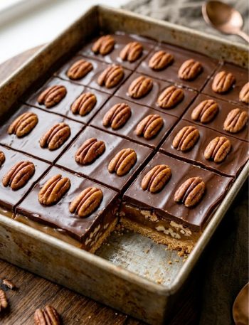 A pan of turtle bars with glossy chocolate topping and whole pecan halves pressed in showing the gooey caramel and graham cracker crust layers