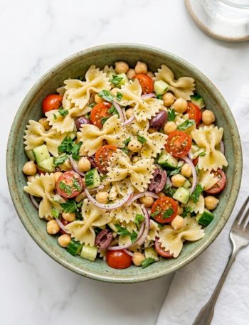 A large ceramic bowl filled with vegan Greek pasta salad made with farfalle pasta, chickpeas, cherry tomatoes, cucumber, kalamata olives, and red onion tossed in a bold lemon herb dressing