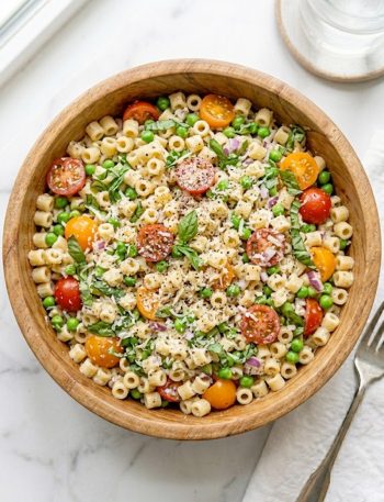 A large wooden bowl filled with vibrant ditalini pasta salad loaded with halved cherry tomatoes, green peas, fresh basil leaves, diced red onion, and Parmesan cheese tossed in a light creamy Italian dressing on a marble surface