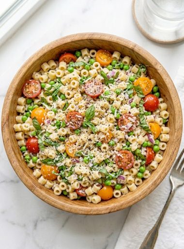 A large wooden bowl filled with vibrant ditalini pasta salad loaded with halved cherry tomatoes, green peas, fresh basil leaves, diced red onion, and Parmesan cheese tossed in a light creamy Italian dressing on a marble surface