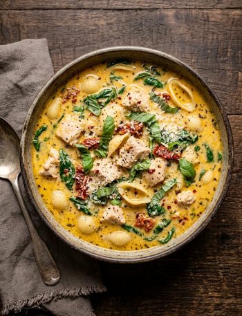 A large bowl of creamy Tuscan chicken soup with tender chicken chunks, shell pasta, wilted spinach, and sun-dried tomatoes in a golden cream broth, photographed on a dark rustic wooden surface