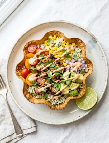 A crispy baked tortilla bowl filled with seasoned grilled chicken, cilantro lime rice, sweet corn, fresh tomatoes, red onion, avocado chunks, and a creamy chipotle drizzle on a white surface