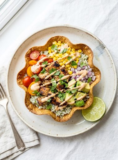 A crispy baked tortilla bowl filled with seasoned grilled chicken, cilantro lime rice, sweet corn, fresh tomatoes, red onion, avocado chunks, and a creamy chipotle drizzle on a white surface
