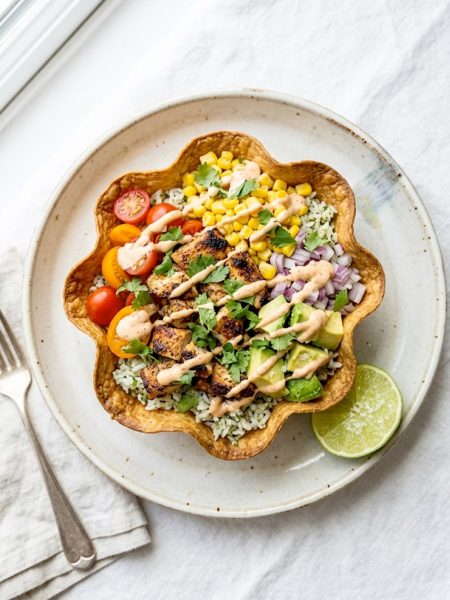 A crispy baked tortilla bowl filled with seasoned grilled chicken, cilantro lime rice, sweet corn, fresh tomatoes, red onion, avocado chunks, and a creamy chipotle drizzle on a white surface