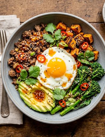 A gray ceramic bowl filled with seasoned ground beef, roasted sweet potato cubes, broccolini, sliced avocado, and a fried egg drizzled with sticky hot honey sauce and garnished with sesame seeds, red chili, and fresh cilantro on a wooden surface