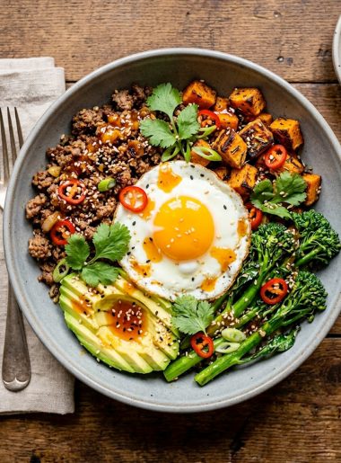 A gray ceramic bowl filled with seasoned ground beef, roasted sweet potato cubes, broccolini, sliced avocado, and a fried egg drizzled with sticky hot honey sauce and garnished with sesame seeds, red chili, and fresh cilantro on a wooden surface