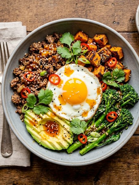 A gray ceramic bowl filled with seasoned ground beef, roasted sweet potato cubes, broccolini, sliced avocado, and a fried egg drizzled with sticky hot honey sauce and garnished with sesame seeds, red chili, and fresh cilantro on a wooden surface